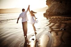 a man and woman walking on the beach with their arms in the air as the sun goes down