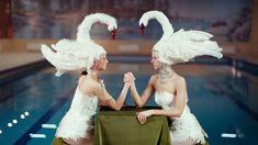 two women sitting at a table in front of a swimming pool with swans on their heads