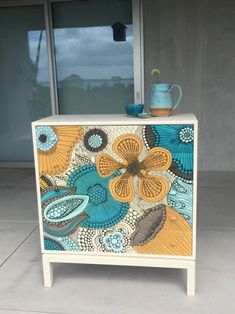 an artisticly painted dresser with a blue vase on it's top and window in the background