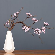 a white vase filled with pink flowers sitting on top of a wooden table next to a gray wall