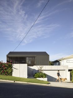 a woman walking her dog on the sidewalk in front of a house with a black roof