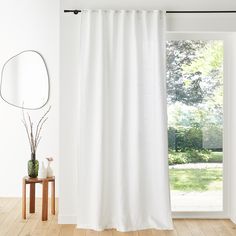 a white curtain hanging in front of a window with a mirror and vase on the floor