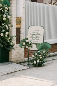 white flowers and greenery are placed in front of a sign on the side of a building