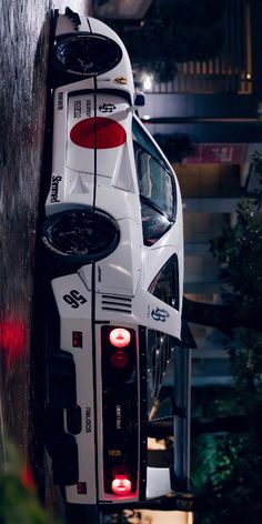 the back end of a white race car with its lights on in front of a building