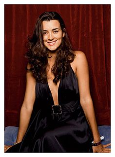 a smiling woman in a black dress sitting on a chair with red curtains behind her