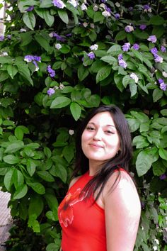 a woman in a red dress standing next to a bush with purple and white flowers