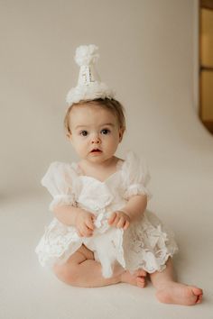 a baby sitting on the floor wearing a white dress and a party hat with ruffles