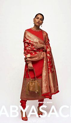 a woman in a red dress and gold shawl is holding a handbag while standing against a white background