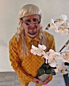 a man with long blonde hair wearing glasses and holding a vase filled with white flowers