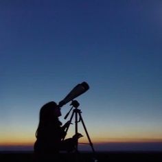 a woman sitting in front of a telescope at night with the sun setting behind her