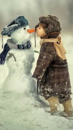 a little boy standing next to a snowman with his nose in the air and wearing a hat