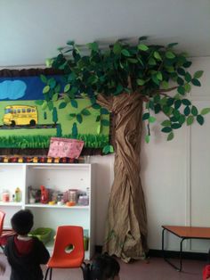 two children are sitting in front of a tree with green leaves on it, and another child is standing next to the bookcase