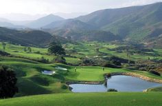 a golf course in the mountains with water and green grass on both sides, surrounded by hills