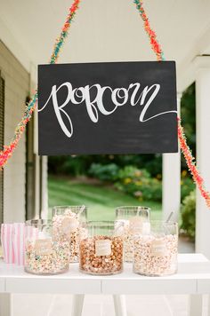 a sign that says popcorn hanging from the side of a table with candy in jars