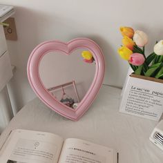 a pink heart shaped mirror sitting on top of a table next to an open book