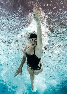 a woman in a black swimsuit swimming under water with her hand up to the air