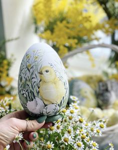 a hand holding an easter egg with a chick on it in front of some daisies