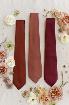 three ties laid out on a table with flowers around them and one is red, the other maroon