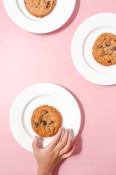 three white plates with chocolate chip cookies on them, one being held by a person's hand