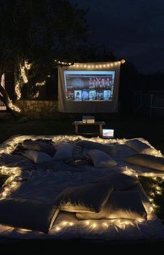 an outdoor movie screen is lit up at night with lights on the ground and pillows all around it