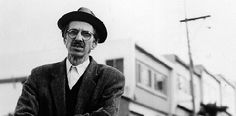 an old man wearing glasses and a hat standing in front of a building on the street