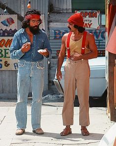 two people standing on the sidewalk eating food and one is wearing a red bandana