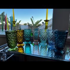 many different colored glass cups on a table near a window with palm trees in the background