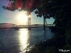 the sun shines brightly behind a bridge over water with trees in front of it