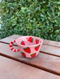 a pink bowl with strawberries on it sitting on a wooden table next to some bushes