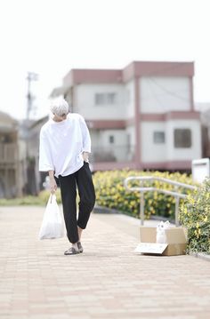 an older woman walking down a sidewalk carrying a white bag and wearing black pants with her foot in the air