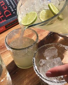 two glasses filled with ice and limes next to a book on a wooden table