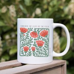 a white coffee mug sitting on top of a wooden table next to some bushes and flowers