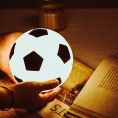 a person holding a lit up soccer ball on top of a table next to an open book