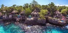 an island with thatched huts on the rocks and people swimming in the blue water