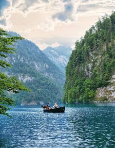 two people in a small boat on a lake surrounded by trees and mountains under a cloudy sky