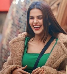 a smiling woman in a green top and brown coat standing on the street with her arms crossed