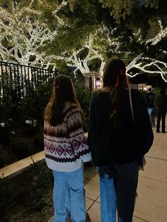 two people walking down a sidewalk with christmas lights on the trees