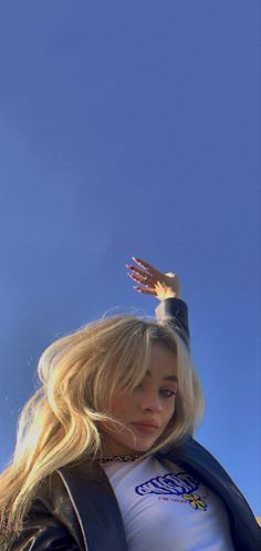 a woman with long blonde hair wearing a white shirt and black leather jacket standing in front of a blue sky