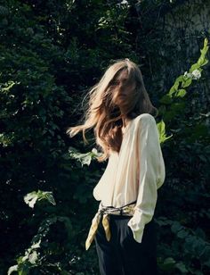 a woman is standing in front of some trees and bushes with her hair blowing in the wind