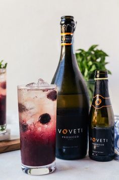 two bottles of wine next to a glass filled with ice and berry drink on a table