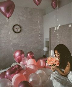 a woman sitting on a bed with balloons floating in the air above her and holding a bouquet of flowers