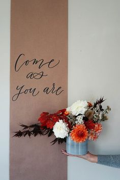 someone holding a vase with flowers in front of a sign that says come as you are