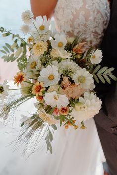 a bride and groom holding a bouquet of white and orange flowers in front of a body of water