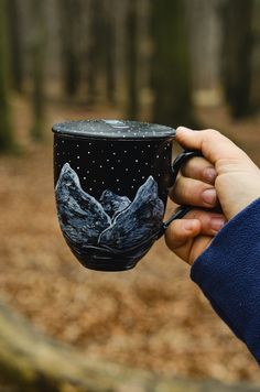 a hand holding a black coffee cup with mountains painted on the side and stars in the sky