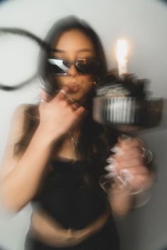 a woman wearing sunglasses holding a cupcake with a lit candle in her hand and looking at the camera