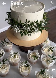 a cake with white frosting surrounded by cupcakes and greenery on a table