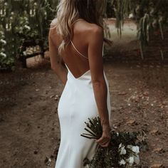 the back of a woman in a white wedding dress holding a bouquet of greenery