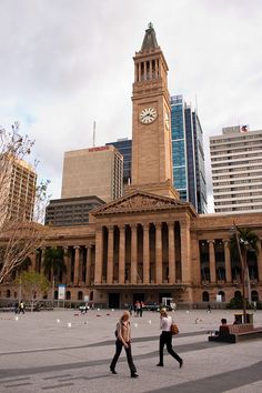 two people walking in front of a large building with a clock on it's tower