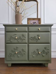 a green dresser with gold knobs and flowers on the top, in front of a mirror