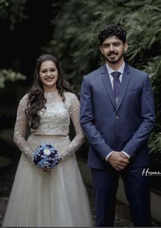 a man and woman standing next to each other in front of some trees with blue flowers